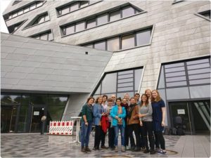 Gruppe von ca. 15 Menschen vor großem grauen Gebäude mit schiefen Fenstern