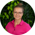 Jana Mätz in front of dark green plant background, blond hair made into bun, pink polo shirt, medium brown glasses, smiling